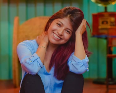 A smiling young woman with dyed hair wearing a light blue shirt in a cozy room.