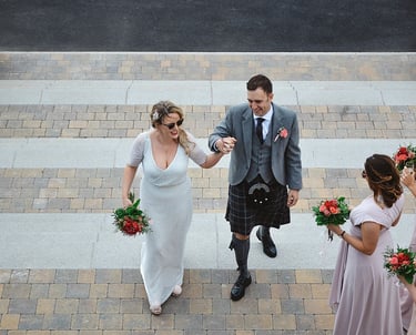 A smiling bride in a white dress and groom in a traditional Scottish kilt walk up stone stairs.