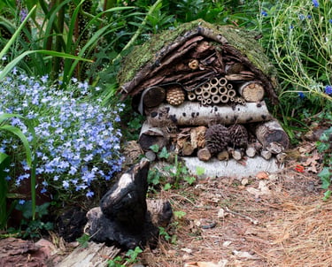 Handmade wooden insect hotel in a lush garden with blue wildflowers and a moss-covered roof.
