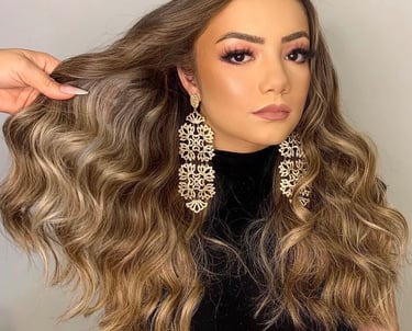 Woman with long wavy balayage hair, glamorous pink eyeshadow, and gold statement chandelier earrings.