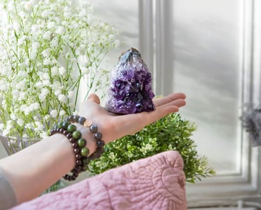 A photo of a hand holding a purple Amethyst cluster crystal with some white flowers