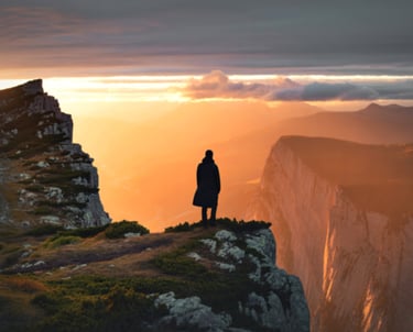 A lone person in a dark coat stands on a mountain cliff overlooking a vast, sunlit valley with rugged peaks at sunset