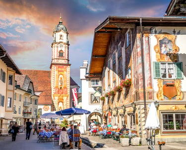 Charming Mittenwald street scene with Lüftlmalerei murals, outdoor cafes, and the St. Peter and Paul church tower.