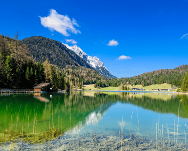 Scenic view of a crystal clear alpine lake in the mountains with a rustic wooden hut and pine forest.