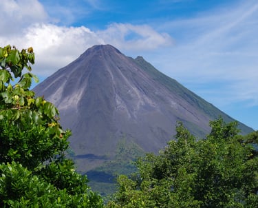 Arenal Volcano in Costa Rica with lush green tropical trees and a blue sky with white clouds.