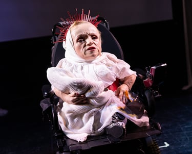 A woman of short stature wearing a white dress and crown of thorns in an electric wheelchair