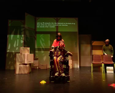 A white electric wheelchair user with a white person balanced on the back, and a BSL interpreter