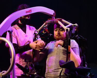 A woman in pink scrubs helps an electric wheelchair user into a hoist and fluffy handcuffs