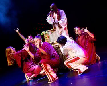 A black person in white robes, with a group of people in red and white at their feet