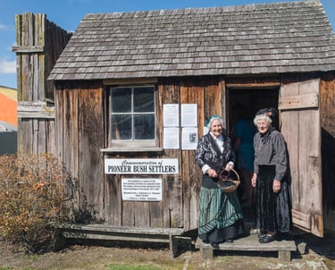 two women in period costumes standing in front of a traditional Pioneer Bush Hut
