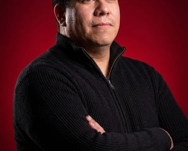 Professional portrait of a middle-aged Hispanic man in a black sweater against a red background.