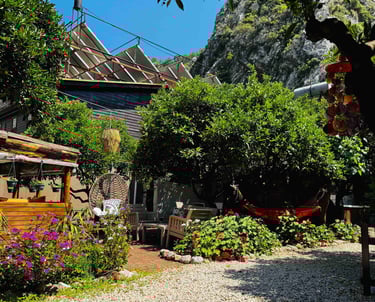 Olympos Kakule Evleri’nin bahçesi; çiçeklerle süslenmiş oturma alanı, hamak ve yemyeşil ağaçlar.