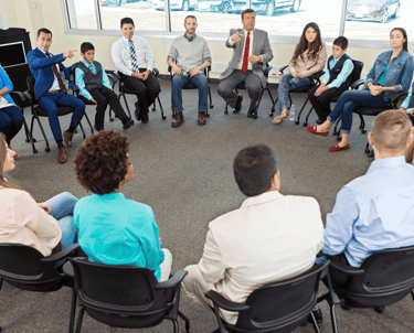 CIlvēki ofisā sēž aplī ar diviem diskusijas vadītājiem