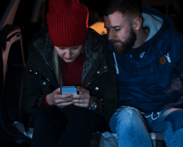 Vīrietis un sieviete sēž uz bagāžnieka vakarā un skatās telefonā