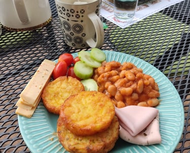 A hearty breakfast plate with hash browns, baked beans, ham, and fresh vegetables on an outdoor patio table.