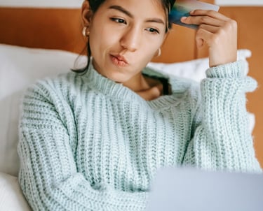 A thoughtful woman using a credit card and laptop for online shopping while sitting in bed.