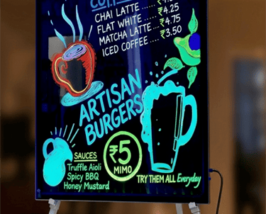 Illuminated neon LED writing board displaying cafe menu specials with colorful fluorescent markers.