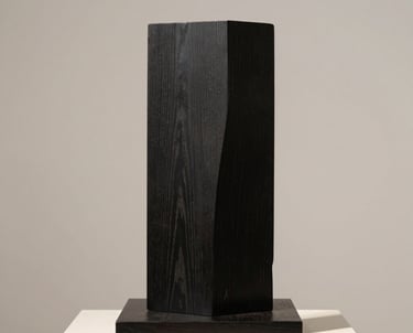 Minimalist photography of a black wooden sculpture on a pedestal against an off-white wall. French gallery style.