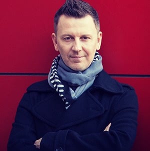 Portrait of a John Taylor in a navy pea coat and scarf against a vibrant red wall.