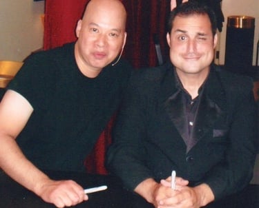 A smiling man in a black suit sitting next to a man in a t-shirt at a meet and greet event.