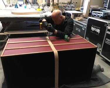 Ray Lum uses a power drill to assemble a custom wooden magic illusion trunk in a workshop.