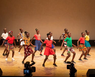 A wide angle shot of a vibrant African / Pan-African youth talent show. A group of kids is performing a choreographed dance on a stage under warm yellow lights, with a professional camera crew in the foreground.