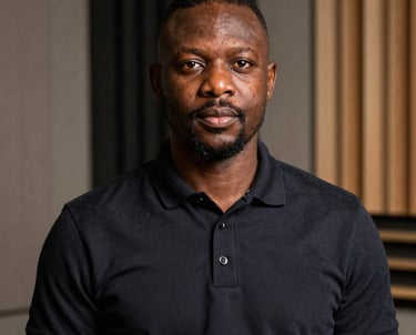 A professional portrait of an African / Pan-African man in a dark polo shirt, standing in front of a background of acoustic studio panels.