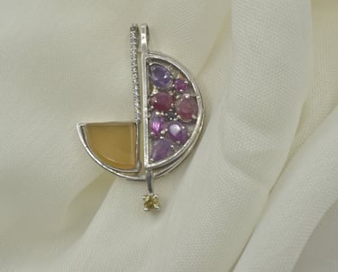 A silver geometric pendant featuring amethyst, ruby, and citrine gemstones on a white fabric background.