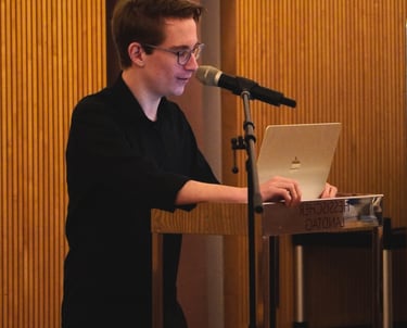 Junger Mann mit Brille hält Vortrag am Laptop und spricht in Mikrofon auf Bühne – Rede