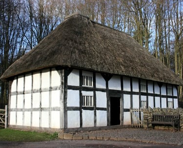 Historic timber-framed cottage with a thick thatched roof situated in a woodland setting.