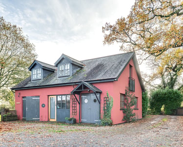 Red country cottage with grey barn doors and dormer windows surrounded by autumn trees.