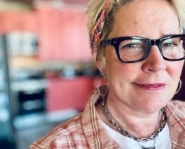 White woman in chunky black glasses and a plaid shirt, wearing a bandana in her hair.
