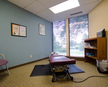 Clean chiropractic exam room with a treatment table, a bookcase of supplies, and a single chair.