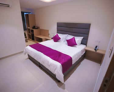 Modern hotel room with a queen bed, grey upholstered headboard, and purple decorative pillows and runner.