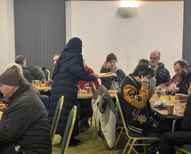 Diverse group of people gathering for a community meal event indoors at long dining tables.