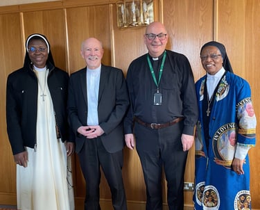 Four religious figures, including nuns and priests, stand together in a formal room with wood paneling, smiling warmly.