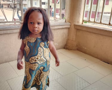 A young girl wearing a blue and yellow patterned Ankara dress walks on a tiled balcony.