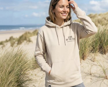 Smiling woman wearing a beige hoodie on a sandy beach with grassy dunes in the background.