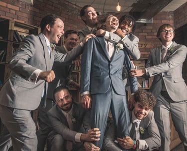 Groomsmen in grey suits laughing and lifting the groom in a blue suit during wedding celebrations.