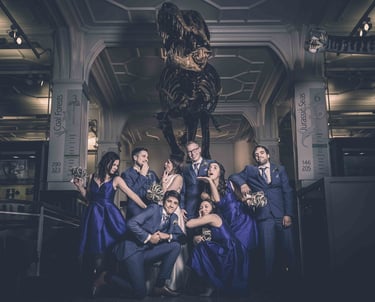 Wedding party in blue attire posing playfully beneath a Tyrannosaurus rex skeleton at a museum venue.