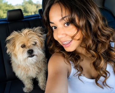 A smiling young woman with curly hair takes a car selfie with her small, fluffy tan dog.
