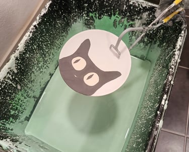 A ceramic plate with a hand-painted black cat being dipped into a bucket of green pottery glaze.
