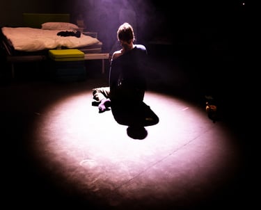 Tatum sits on the floor looking away from the audience, bathed in a spotlight facing their bed