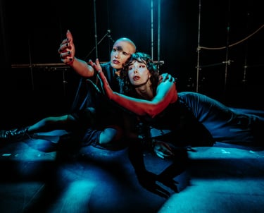 Ila and Coda, bathed in blue light and shadow reach their arms up together, their gazes meeting