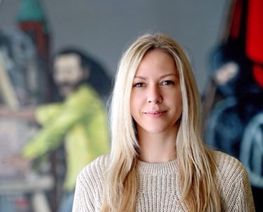 Rikke Lydeking in a beige knit sweater smiling in an art studio with paintings in the background.