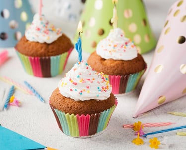 Birthday cupcakes with white frosting, sprinkles, and a lit candle next to party hats.