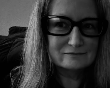 A black and white close-up portrait of a woman with long hair wearing thick black framed glasses.