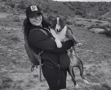 Mujer sonriente con mochila caminando al aire libre mientras sostiene a su Bull Terrier atigrado.