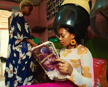 a woman in a colorful dress is reading a magazine while sitting under dryer in a hairdresser's shop 