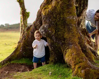 Mother watching toddler son explore around ancient tree trunk during natural family PHOTOSHOOT